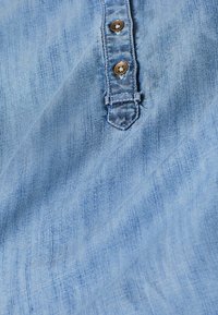 Jasnoniebieska tkanina o subtelnej teksturze, z zapięciem na guziki i zaokrąglonym dolnym brzegiem, z trzema brązowymi guzikami na środku.