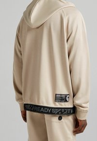 Beige mikina s kapucí a odpovídající kalhoty s černým elastickým pasem, na kterých je bílý a šedý text, nošené osobou zezadu.
