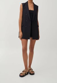 Black sleeveless tailored blazer with double-breasted buttons, paired with a black top, distressed shorts, and tan platform sandals.
