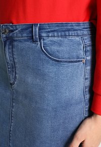 Gonna di denim azzurro chiaro con taglio diritto, tasca frontale e dettaglio con bottone argento. Indossata con una maglia rossa, che mette in risalto il suo design informale.