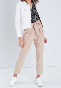 Veste en denim blanche, blouse noire à motifs, pantalon beige sur mesure avec ceinture nouée, et baskets blanches, mettant en valeur une coupe structurée et un style décontracté.