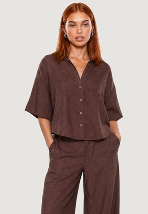 Femme aux cheveux roux portant une chemise marron ample boutonnée avec des manches trois quarts et un pantalon large assorti, mains dans les poches.