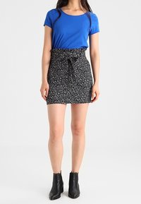 Blaues kurzärmliges T-Shirt, kombiniert mit einem schwarzen Minirock mit floralen Mustern und gebundenem Bund. Die Modell trägt schwarze Stiefeletten.