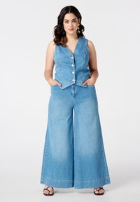 Model in een chambray cropped vest en wijde jeans, met handen in de zakken, staat zelfverzekerd tegen een eenvoudige witte achtergrond.