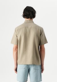 Chemise à manches courtes en matériau texturé beige avec une emmanchure et un col dans le dos. Motif gaufré sur l'ensemble, associée à un short bleu clair.