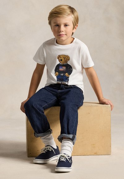 Młody chłopiec o blond włosach siedzi na drewnianym bloku, ubrany w białą koszulkę z grafiką niedźwiedzia, ciemne dżinsy, białe skarpetki i granatowe trampki.