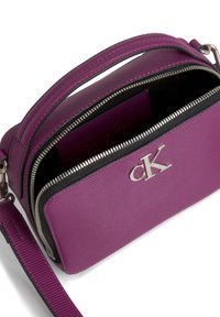 Fioletowa torebka crossbody Calvin Klein z przednią kieszenią na zamek, srebrnym logo cK i regulowanym paskiem, pokazana częściowo otwarta.
