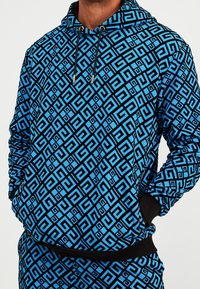 Modrá mikina s geometrickým černým vzorem. Šňůrky se zlatými detaily, žebrované manžety a lem. Měkká látka s volným střihem.