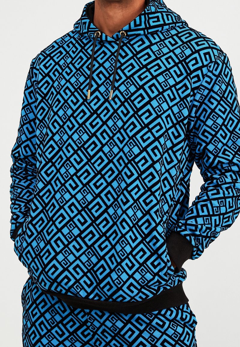 Modrá mikina s geometrickým černým vzorem. Šňůrky se zlatými detaily, žebrované manžety a lem. Měkká látka s volným střihem.