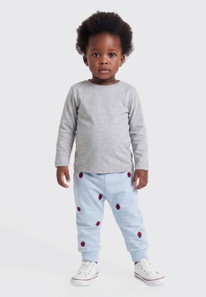Jeune enfant debout, portant une chemise à manches longues gris clair, un pantalon bleu clair avec des motifs de fraises rouges et des baskets blanches.