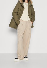 Olivově zelená prošívaná parka s kapucí lemovanou kožešinou, krémový pletený svetr, béžové kalhoty se sklady a náprsními kapsami, a černo-bílé tenisky.