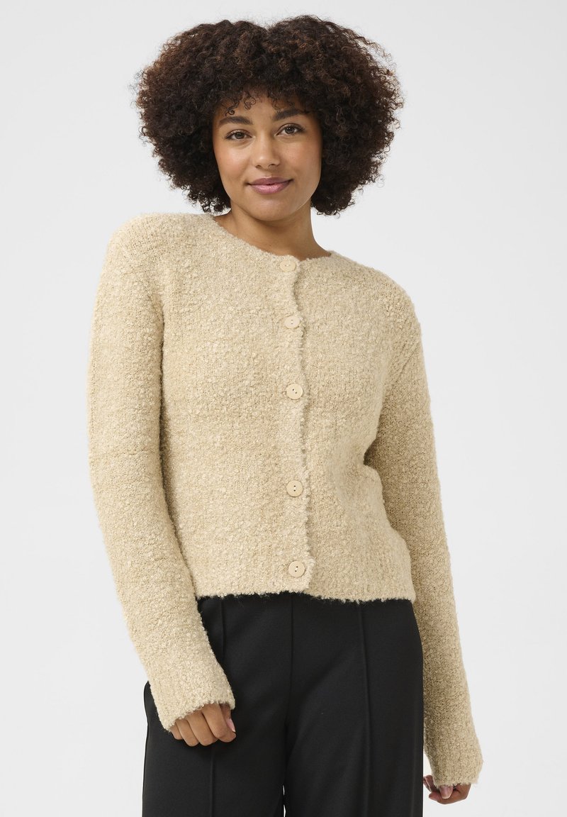 Texturerad beige kofta med långa ärmar, rund hals och fem ljusa träknappar framtill. Bärs med svarta byxor.