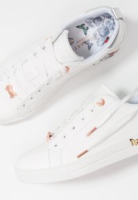 Baskets basses blanches avec œillets en or rose et détails nœuds, motif papillon sur la semelle et le côté, lacets blancs sur fond blanc.