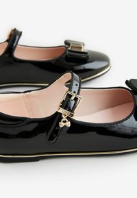 Zapatos Mary Jane de charol negro con un lazo y detalles de hebilla dorada, con un suave forro interno rosa y un borde dorado en la suela.