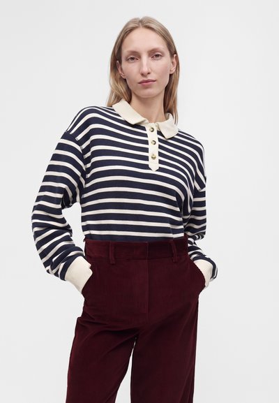 Femme blonde debout, mains dans les poches, portant un polo à manches longues rayé bleu marine et blanc et un pantalon en velours côtelé bordeaux taille haute.