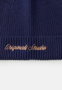 Jūras zils adīts cimds ar ribotu tekstūru un dekoratīvu vara krāsas izšuvumu "Originals Studio" priekšpusē.