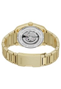 Guldfarvet rustfrit stål ur med en flad, oktagonal urkasse, automatisk urværk synligt gennem en gennemsigtig bagside, og et tekstureret ledarmbånd.