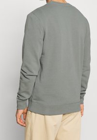 Mężczyzna w jasnoszarej bluzie z długim rękawem i beżowych spodniach, pokazany z tyłu na tle białym.