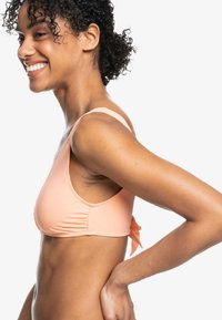 Perzikkleurige bikini-top met dunne bandjes, voorzien van zijkanten met plooien en een gestrikt accent aan de achterkant. Glad materiaal en minimalistisch ontwerp.