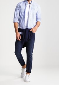 Herroutfit med en ljusblå rutig skjorta med knappar, vit T-shirt, marinblå tröja knuten i midjan, mörka jeans och vita sneakers.