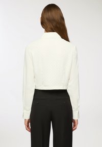 Blusa bianca cropped con maniche lunghe, caratterizzata da un sottile motivo a pois, polsini con bottoni e colletto classico, abbinata a pantaloni neri.