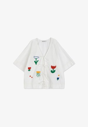 Witte cropped top met een losse pasvorm, versierd met bloemenborduurwerk in rood, blauw, geel en groen. De strikjes aan de voorkant creëren een V-halslijn.