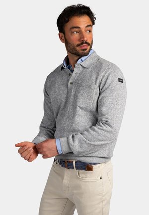 Man met een grijze fleece trui over een blauw overhemd met kraag, beige broek en een blauwe geweven riem, die zijn mouw afstelt terwijl hij naar rechts kijkt.