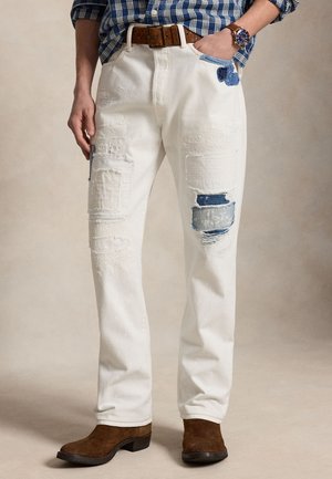 Man draagt witte patchwork jeans met slijtage, bruine suède laarzen, een bruine leren riem en een blauw geruit overhemd met horloge om de pols.