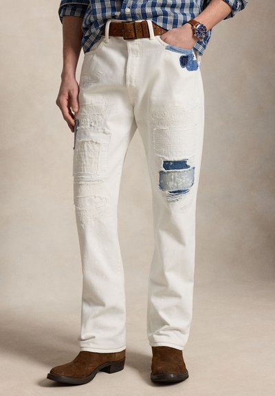 Man som bär vita patchwork slitna jeans, bruna mockastövlar, ett brunt läderbälte och en blå rutig skjorta med klocka på handleden.