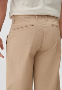 Beige katoenen broek met een gladde textuur, voorzien van twee achterzakken en een aansluitende tailleband. Subtiele stikdetails zijn zichtbaar.