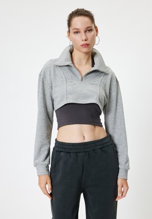 Sudadera gris corta con cuello alto y cierre de cremallera, con mangas largas y costuras planas. Top negro ajustado y pantalones de chándal oscuros abajo.
