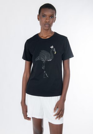 Persona con camiseta negra con un diseño de flamenco con lentejuelas y corona, y pantalones cortos blancos, de pie contra un fondo blanco liso.