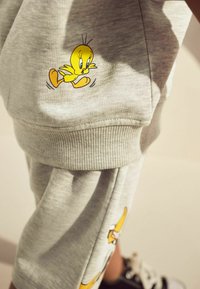 Grå bomullsshorts med en tecknad gul fågelgrafik, ribbade fållar och en slät yta. Detaljer inkluderar ytterligare fågelillustrationer.
