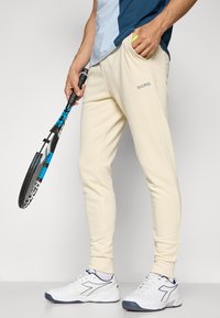 Cream tapered sweatpants with BORG logo, side pockets, and ribbed cuffs, paired with white athletic shoes and holding a tennis racket.