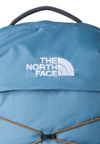 Sininen reppu mustalla kahvalla, valkoinen kirjailtu "The North Face" -logo ja ruskea joustava nyöri etutaskun yli.