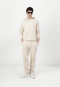 Felpa beige con motivo e cappuccio, abbinata a pantaloni sportivi coordinati. Entrambi presentano una texture morbida e una vestibilità comoda. Sneakers bianche completano il look.