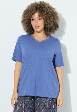 Mujer con cabello rizado que lleva una camiseta azul de cuello en V y manga corta y pantalones estampados en blanco y negro, de pie contra una pared blanca.