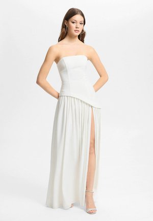 Femme portant une robe blanche sans bretelles avec un corsage asymétrique et une fente haute sur le côté, debout les mains sur les hanches devant un fond uni.