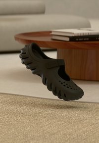 Sabot léger noir avec trous de ventilation et bride réglable, flottant au-dessus d'un tapis beige dans un salon.