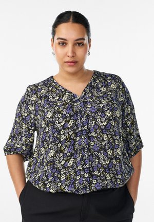 Žena s tmavými vlasmi v voľnej kvetinovej blúzke s fialovými, bielymi a čiernymi kvetmi, stojaca so zbraňami v čiernych nohaviciach.