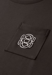 Dunkelbrauner Stoff mit einer aufgenähten Tasche, die ein glänzendes, abstraktes silbernes Metallemblem in der Mitte der Tasche zeigt.