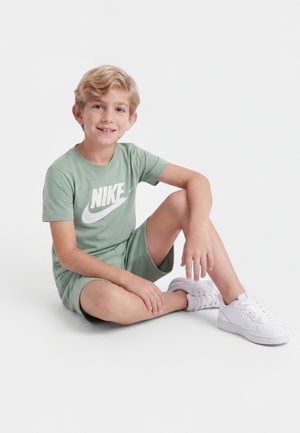 Ξανθό αγόρι καθισμένο με σταυρωμένα πόδια, φοράει πράσινη μπλούζα και σορτς Nike, λευκά αθλητικά παπούτσια Nike, χαμογελά σε λευκό φόντο.