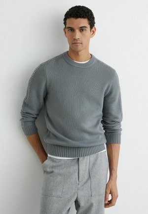 Junger Mann mit kurzen lockigen Haaren, der einen hellgrauen Strickpullover und graue Hosen trägt und vor einer weißen, strukturierten Wand steht.