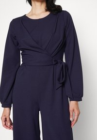 Mörkblå omkrets jumpsuit med en åtsittande midja, långa ärmar och en knutdetalj. Slät textur, har en rund halsringning och plisserad design.