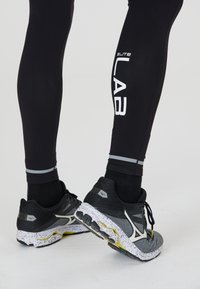 Zwarte sportleggingen met witte tekst "ELITE LAB" en grijze accentlijnen, gecombineerd met zwart-grijze hardloopschoenen met een gespikkelde witte zool.