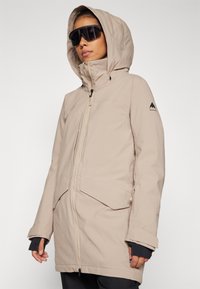 Chaqueta impermeable beige con cremallera frontal, capucha ajustable y grandes bolsillos. Presenta un logo de montaña en la manga y un acabado texturizado.
