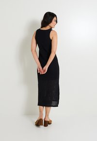 Vestido negro de punto sin mangas, con un patrón texturizado y laterales con aberturas. Largo hasta la mitad de la pantorrilla, combinado con sandalias de tacón grueso en color marrón.