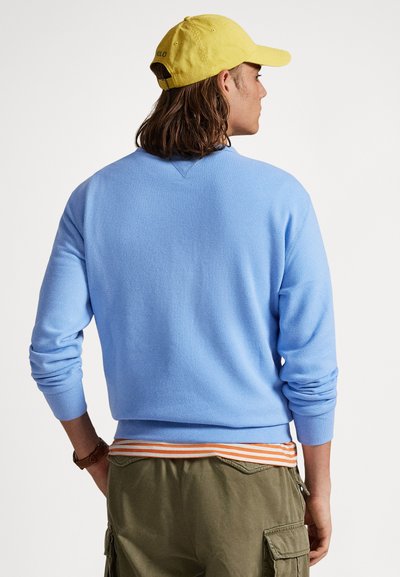 Ανοιχτό μπλε πουλόβερ με στρογγυλή λαιμόκοψη και ριμπ μανσέτες, συνδυασμένο με χακί παντελόνι με πορτοκαλί ριγέ ζώνη μέσης και τσέπες.