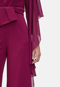 Fuchsia jumpsuit with a deep V-neck, wide sleeves, and a bow accent at the waist, featuring smooth fabric and sheer overlay details.