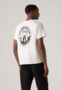 Hvit bomull t-skjorte med et stort svart grafisk design på baksiden, som viser en figur med jeans. Kort ermer og avslappet passform.